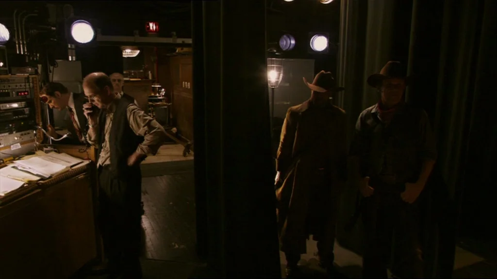 A Prairie Home Companion movie still: wide — A dark technical control area is illuminated by overhead stage lights and desk lamps. Two men dressed…