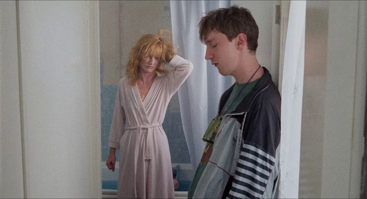Life Is Sweet (1990) movie still: medium — A cramped bathroom contains blue tiled walls and a white shower curtain. A young man stands in a doorway…