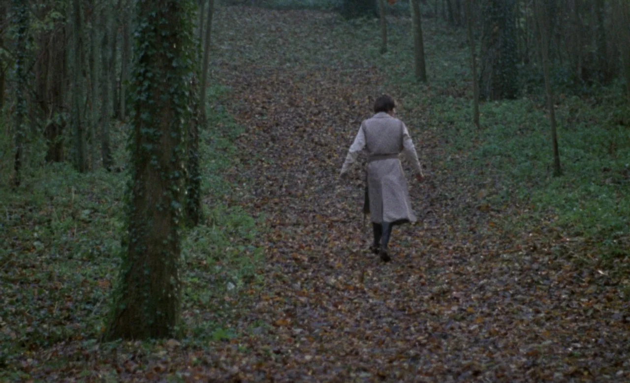Blanche (1971) movie still: wide — A dense woodland path is covered in fallen dead leaves during autumn. A man in a long coat walks away from the…