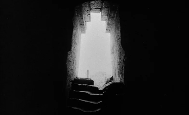 La Jetée (1962) movie still: establishing — A dark stone corridor opens up to bright daylight through a doorway at the end. Rough-hewn stone steps…