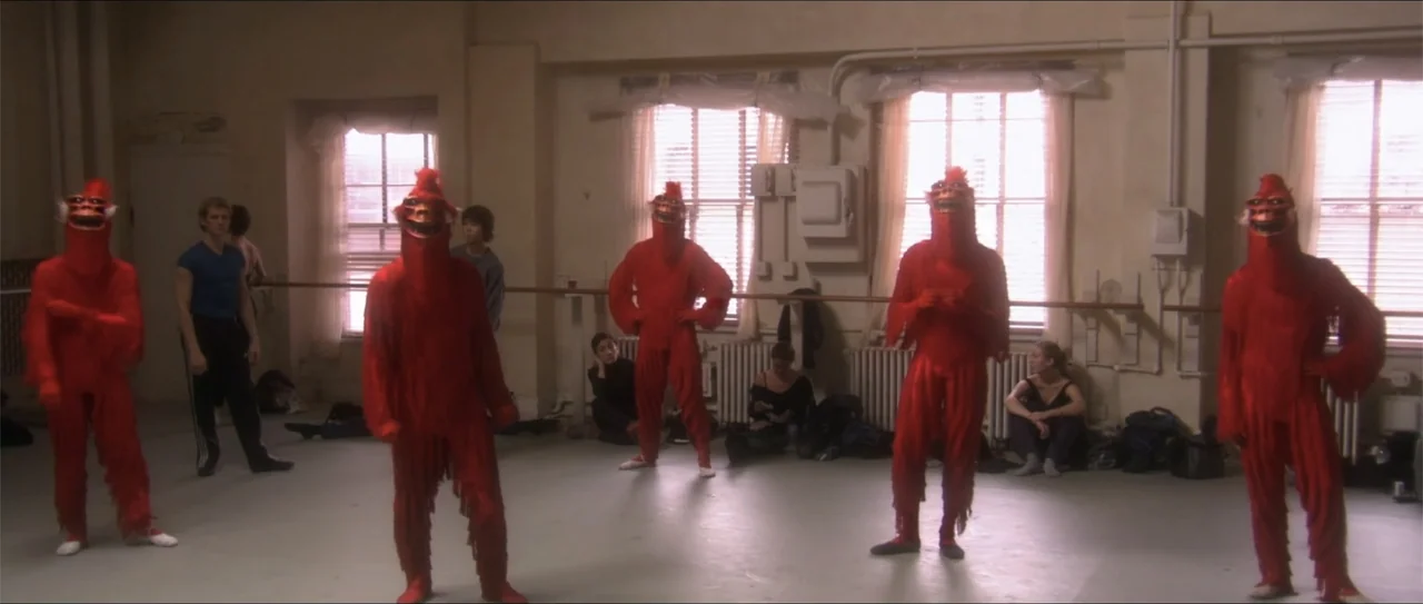 El acto (1989) movie still: fullbody — A dance studio with beige walls, ballet barres, and large windows with closed blinds. Four performers in red…