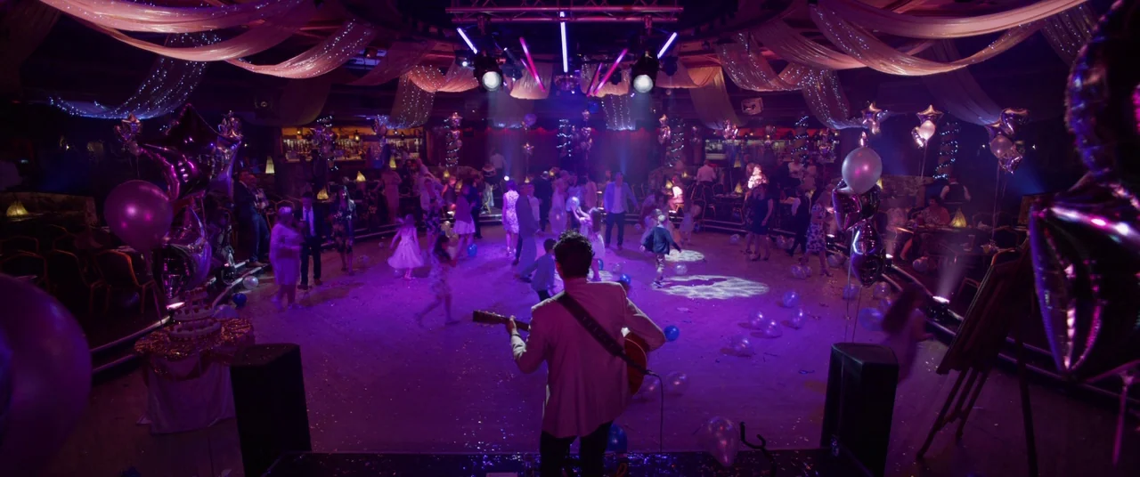 Yesterday (2019) movie still: wide — A spacious dance hall features white fabric swags and scattered balloons across the floor. A musician performs…