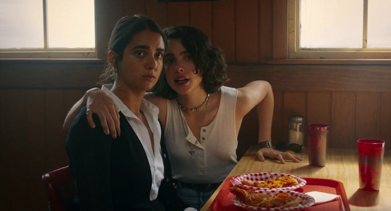 Drive-Away Dykes (2024) movie still: medium — Two young women sit in a wooden-paneled diner booth near bright windows. One woman has her arm around…