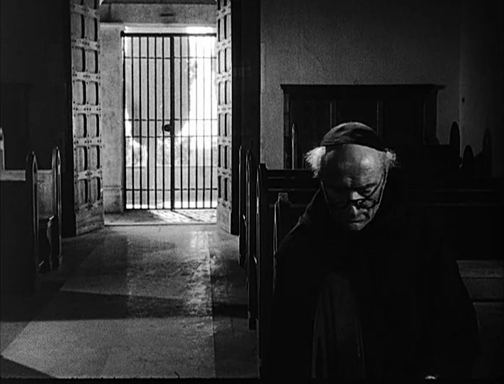 Nights of Cabiria (1957) movie still: medium — A stone interior with wooden pews leads toward a bright iron gate. An elderly monk sits in the…