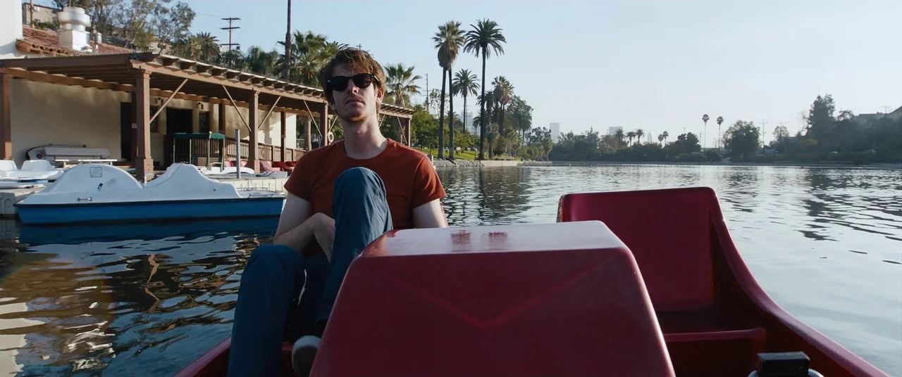 Under The Silver Lake (2018) movie still: medium — A man sits in a red pedal boat on a calm urban lake. Tall palm trees and a shoreline building…
