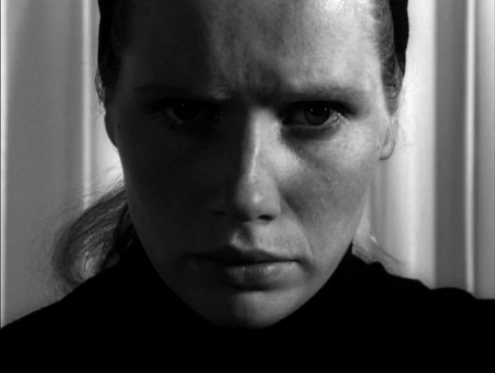 Persona (1966) movie still: closeup — A close portrait shot displays a woman against a plain vertical-striped wall background. The subject maintains…