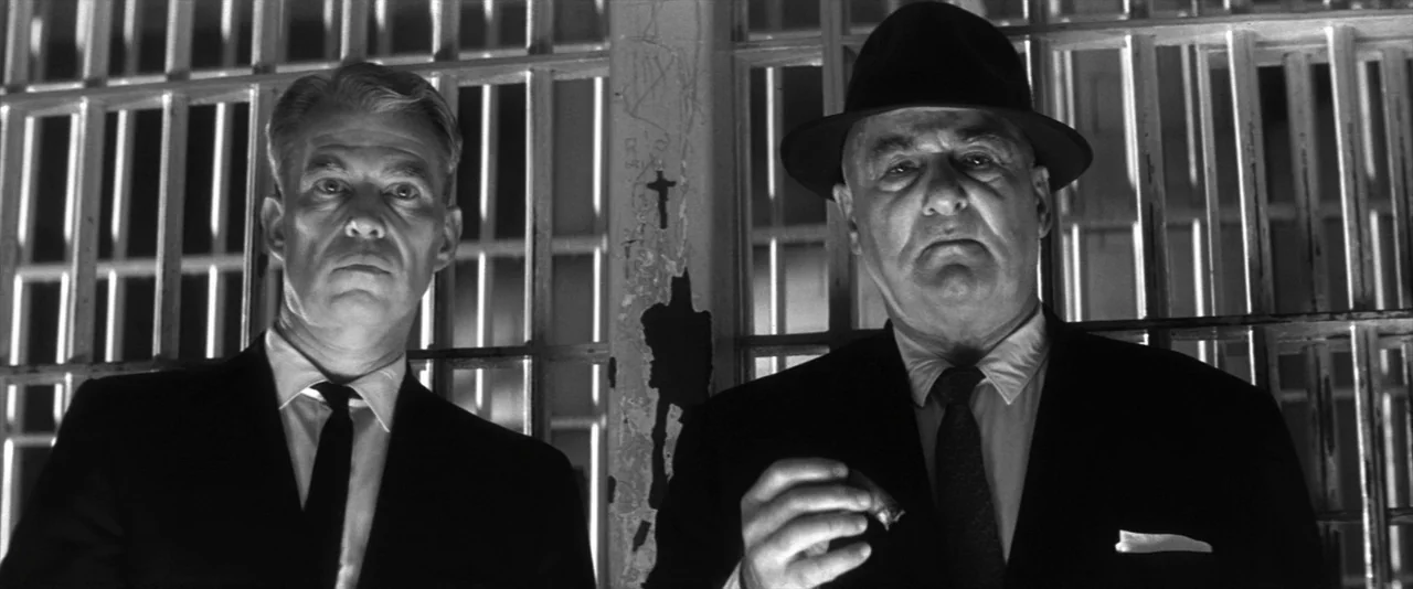 JFK (1991) movie still: medium — A prison cell interior features vertical iron bars in the background behind two men. The men stand together and…