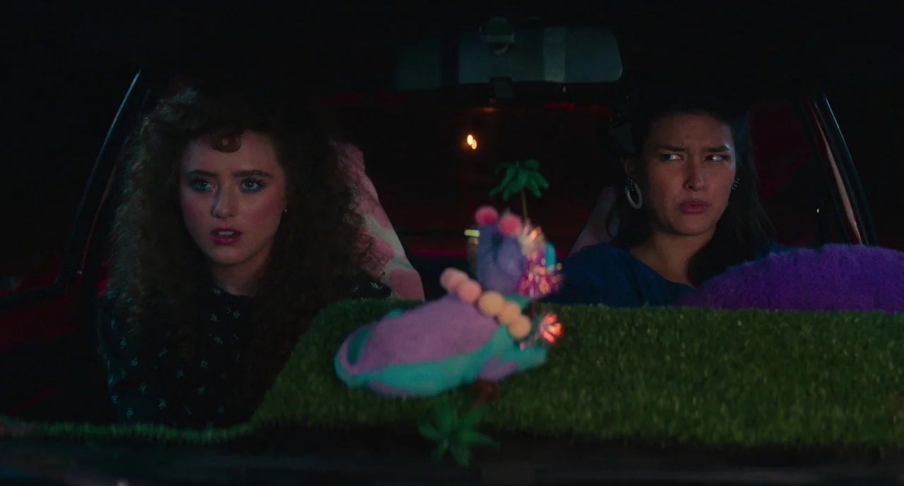 Lisa Frankenstein (2024) movie still: medium — Two women are seated inside a dark car with a grass-covered dashboard surface. A small plush purple…