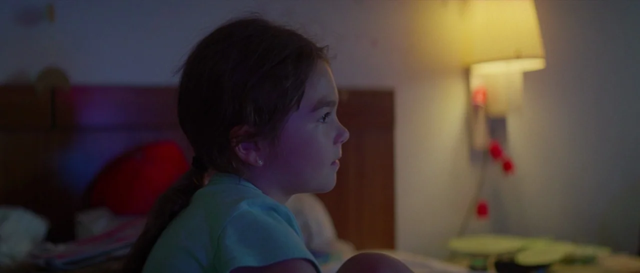 The Florida Project (2017) movie still: medium — A dark hotel room features a wood-paneled headboard and a lit wall-mounted reading lamp. A young…