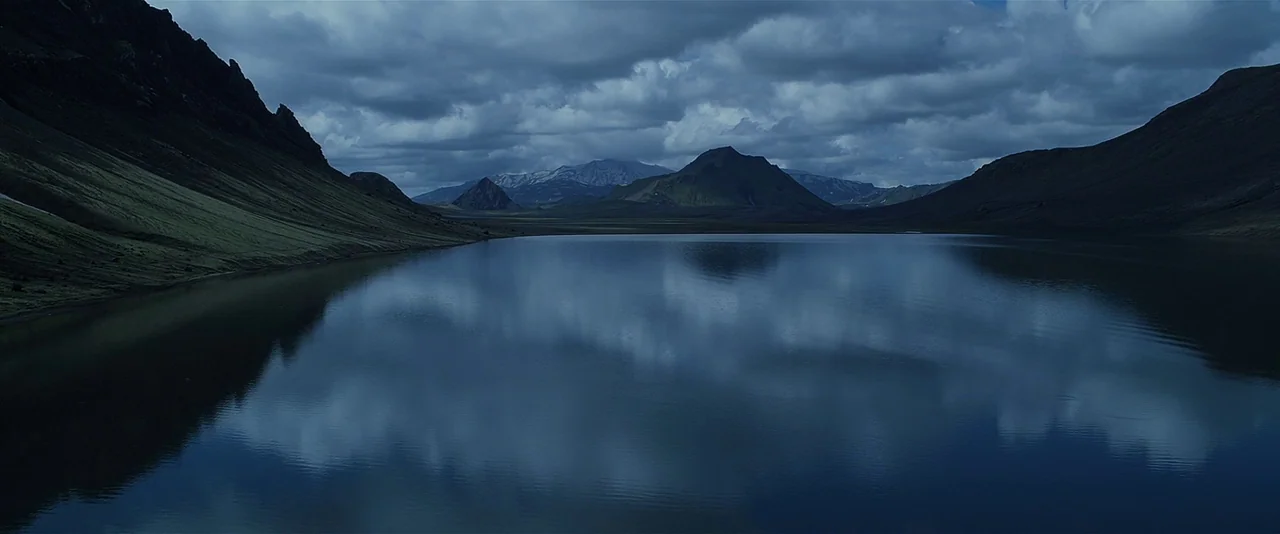 A vast alpine lake sits surrounded by steep, green-sloped mountains under a heavy overcast sky. The water reflects the dark clouds and surrounding terrain in its calm, glassy surface.