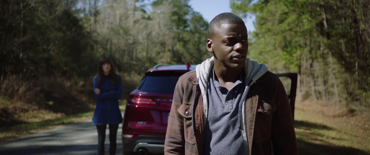Get Out (2017) movie still: medium — A rural paved road cuts through a dense forest with tall pine trees. A man stands in the foreground near a…