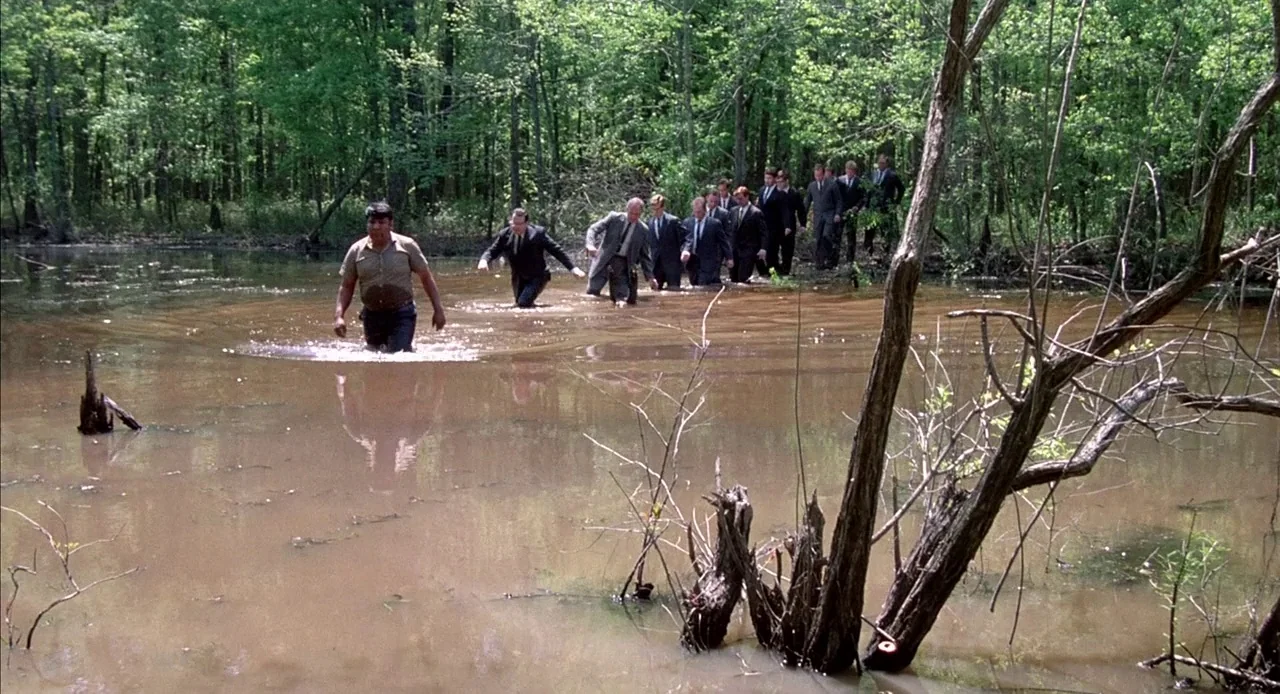 Mississippi Burning (1988) movie still: wide — A group of men in business attire navigate through a shallow, muddy forest swamp. The terrain is…