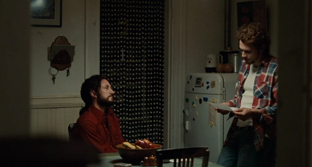 Milk (2008) movie still: medium — An interior domestic space features a table with a fruit bowl and refrigerator. One man stands reading a paper…