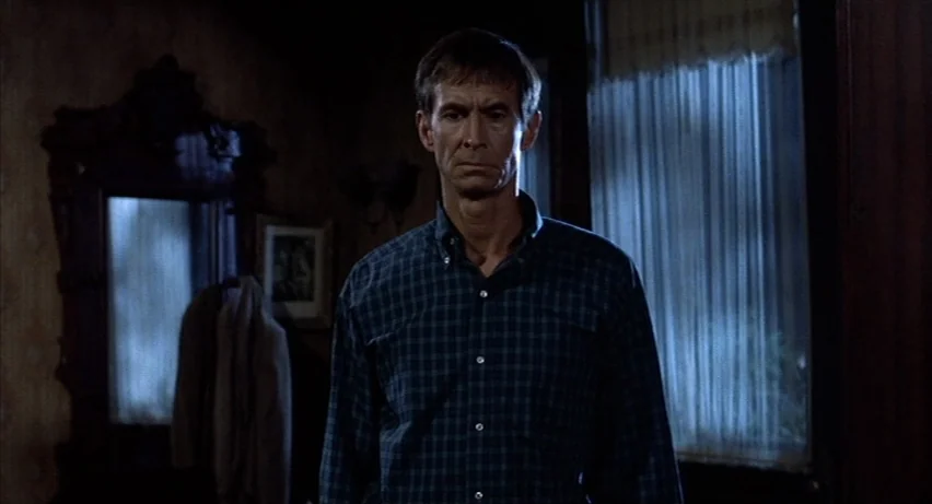 Psycho 2 (1983) movie still: medium — A dimly lit room with dark wooden paneling and sheer window curtains. A middle-aged man stands in the center…