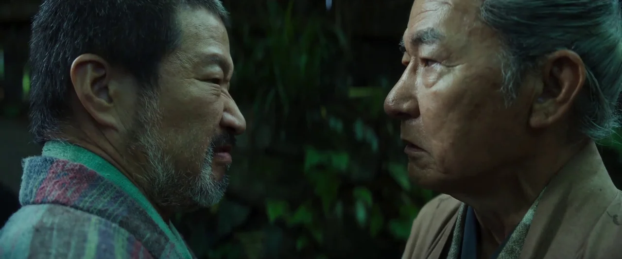 Kubi (2023) movie still: medium — Two men stand closely together against a backdrop of dense, dark green foliage. They face each other in an outdoor…