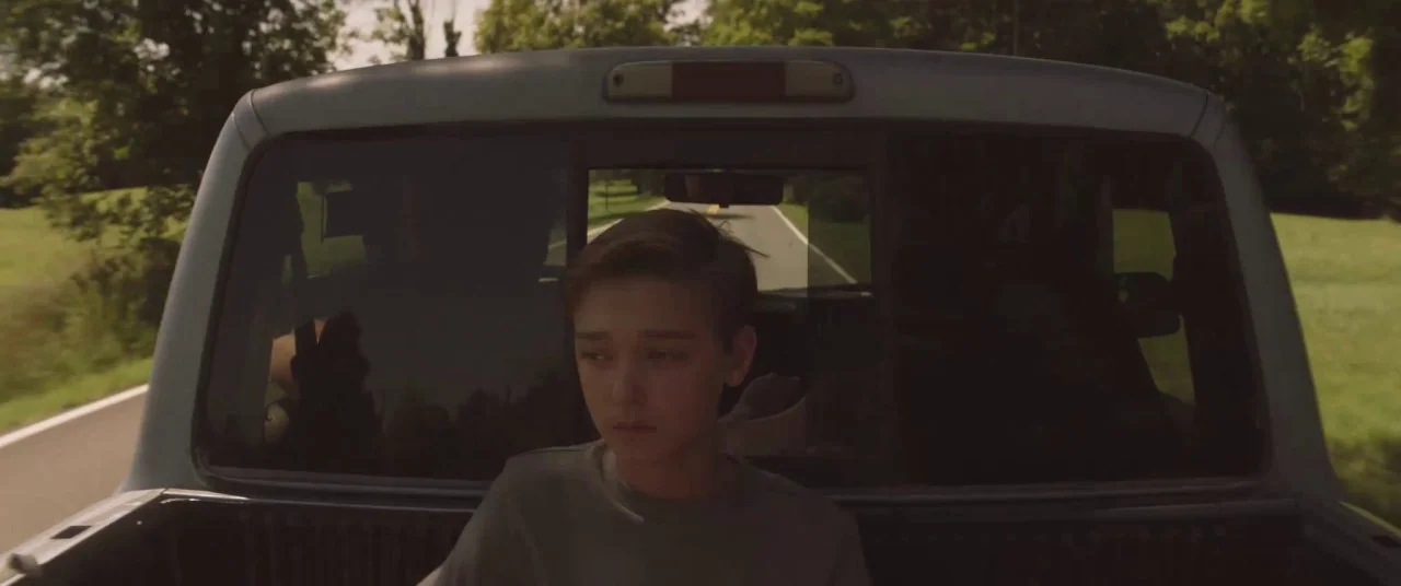 The Strange Ones (2017) movie still: medium — A teenager sits in the back of a truck traveling along a rural road. The truck is in motion with…
