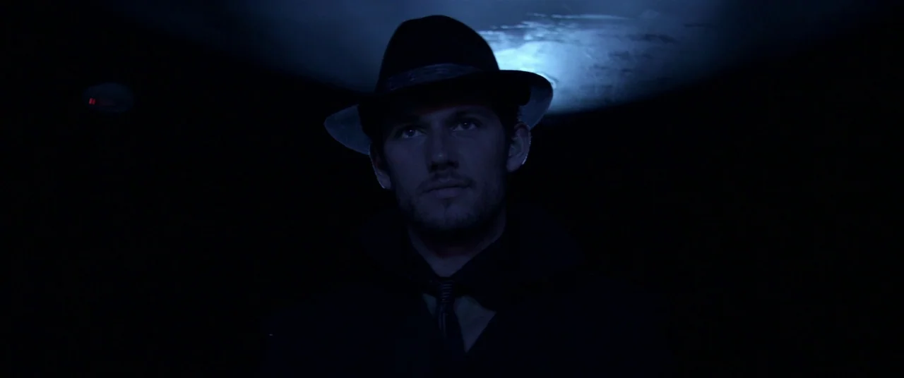Magic Mike (2012) movie still: medium — A man stands in a near-total darkness illuminated by a single overhead cool light. The man wears a fedora…
