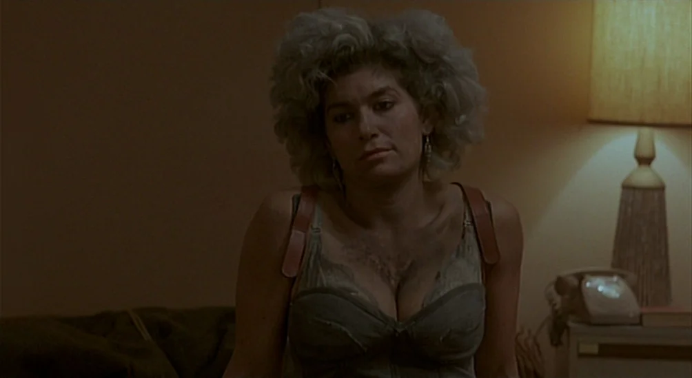 Near Dark (1987) movie still: medium — A dimly lit room with muted walls and a vintage table lamp on a nightstand. A woman with voluminous hair…