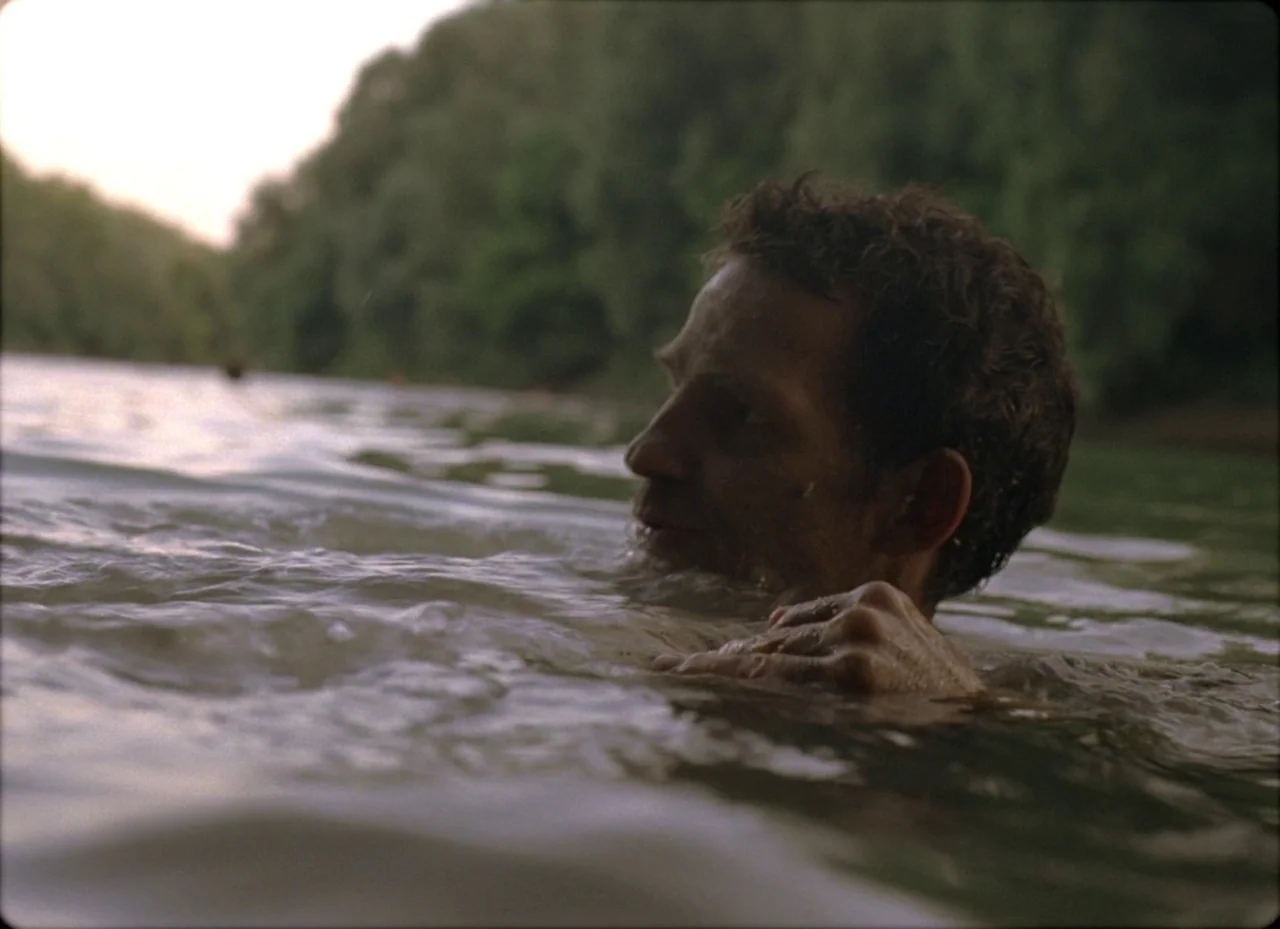 Son of Saul (2015) movie still: medium — A man swims in a calm river with dense green foliage along the bank. The water ripples around his shoulders…
