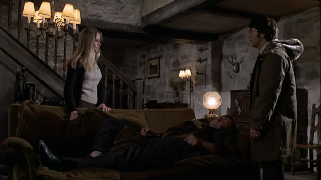 Straw Dogs (1971) movie still: wide — A rustic living room features rough-hewn stone walls, exposed wooden beams, and a heavy chandelier. A woman…