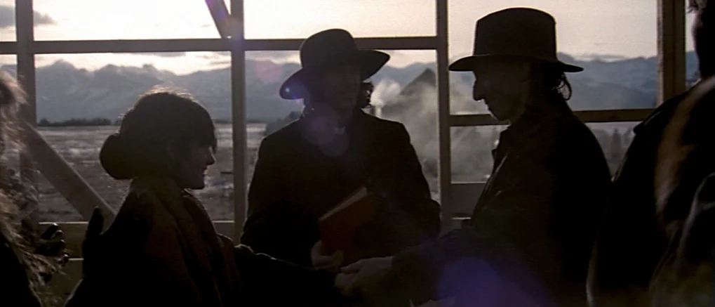 The Claim (2000) movie still: medium — The unfinished frame of a wooden building stands against a wide, mountainous horizon. A group of people in…