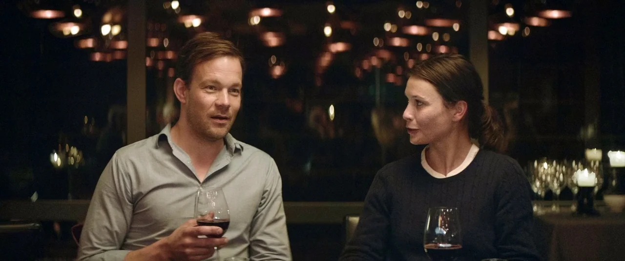 Force Majeure (2014) movie still: medium — A dimly lit upscale restaurant with out-of-focus background lights visible through a window. A man and a…