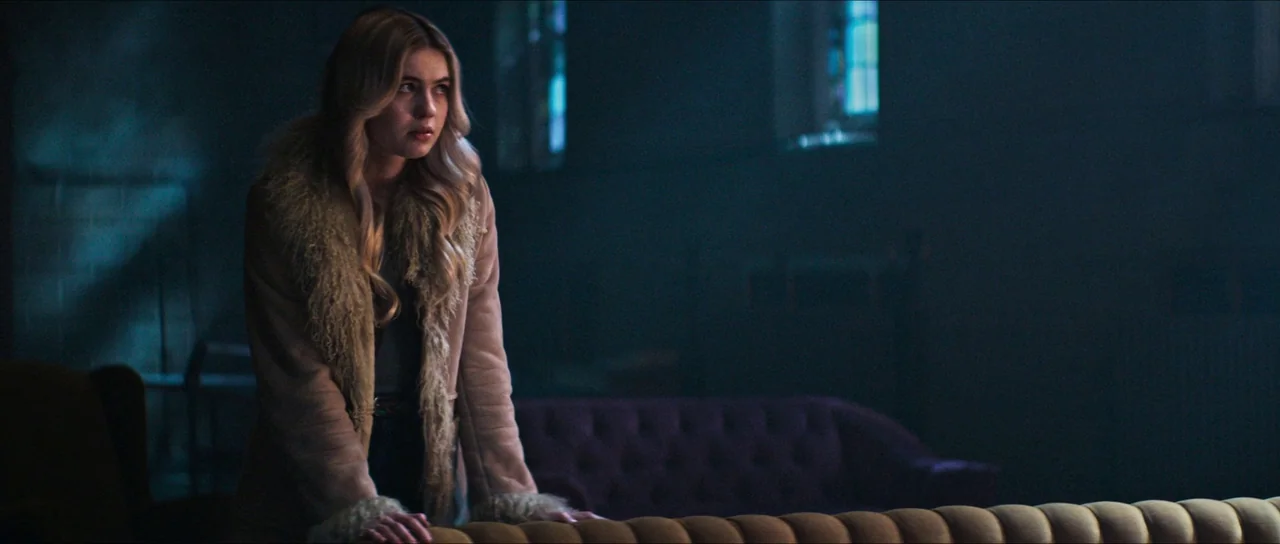 The Sacrifice Game (2023) movie still: medium — A dimly lit lounge with dark walls and a blurred purple sofa in the background. A young woman in a…