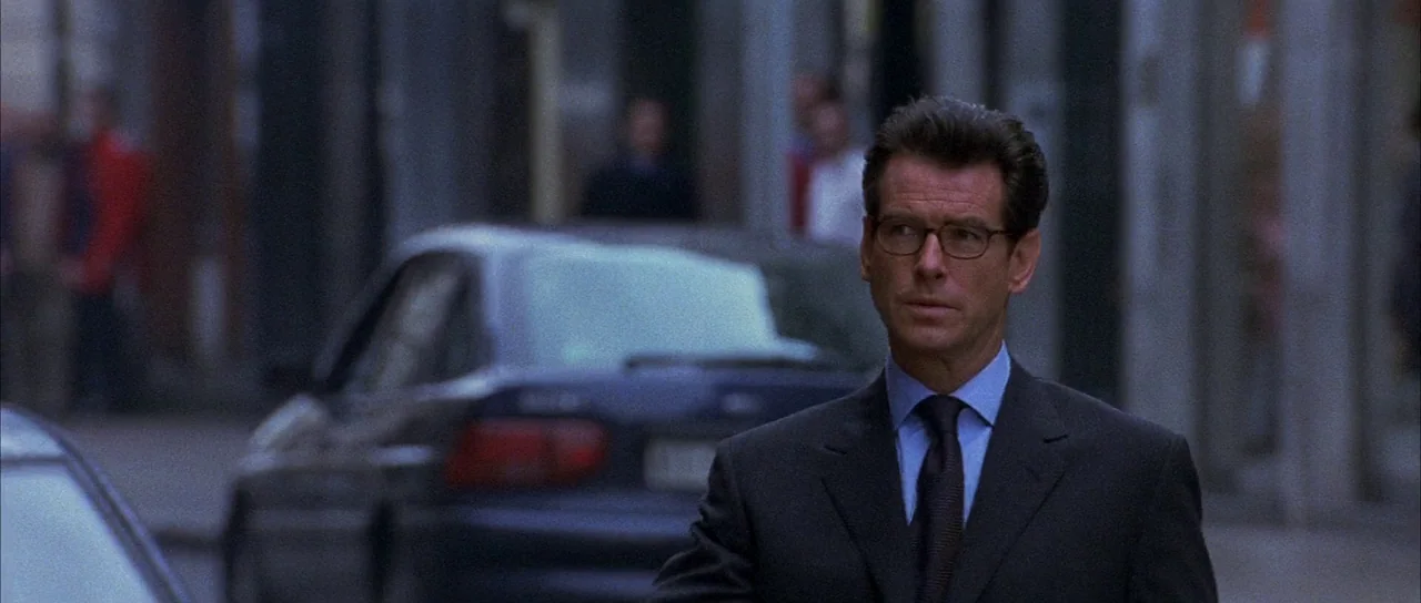 The World is Not Enough (1999) movie still: medium — An urban street with blurry background traffic and blurred figures on a sidewalk. A man in a…