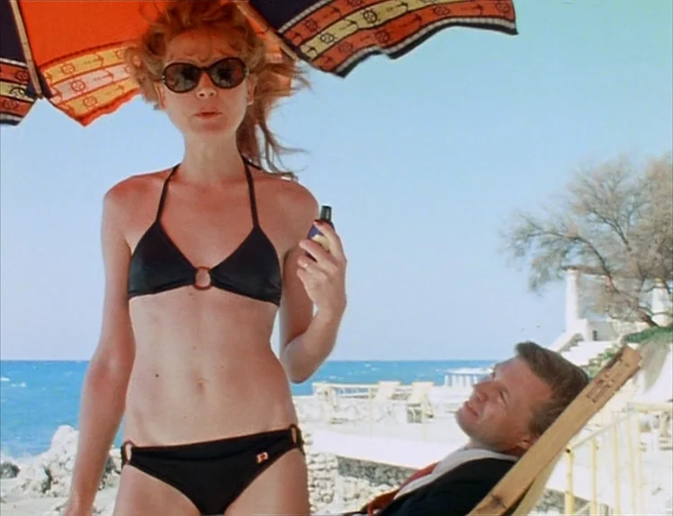 Martha (1974) movie still: medium — A woman stands under a colorful striped umbrella on a coastal stone patio. She holds a small bottle while a man…