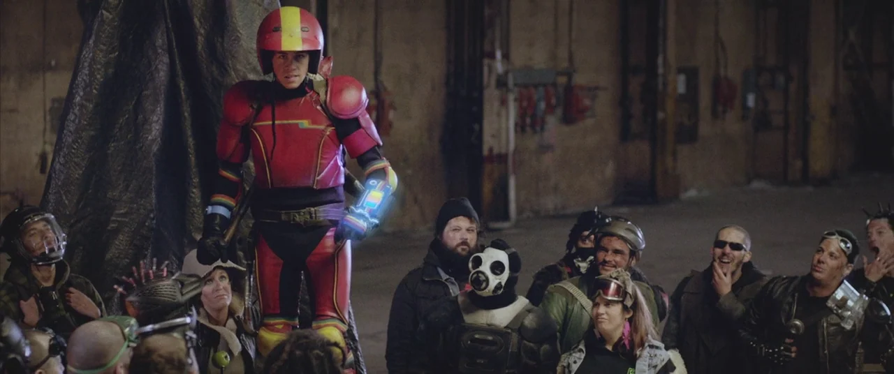 Turbo Kid (2015) movie still: medium — An industrial hall with concrete floors and distant equipment along the walls. A character in a vibrant…