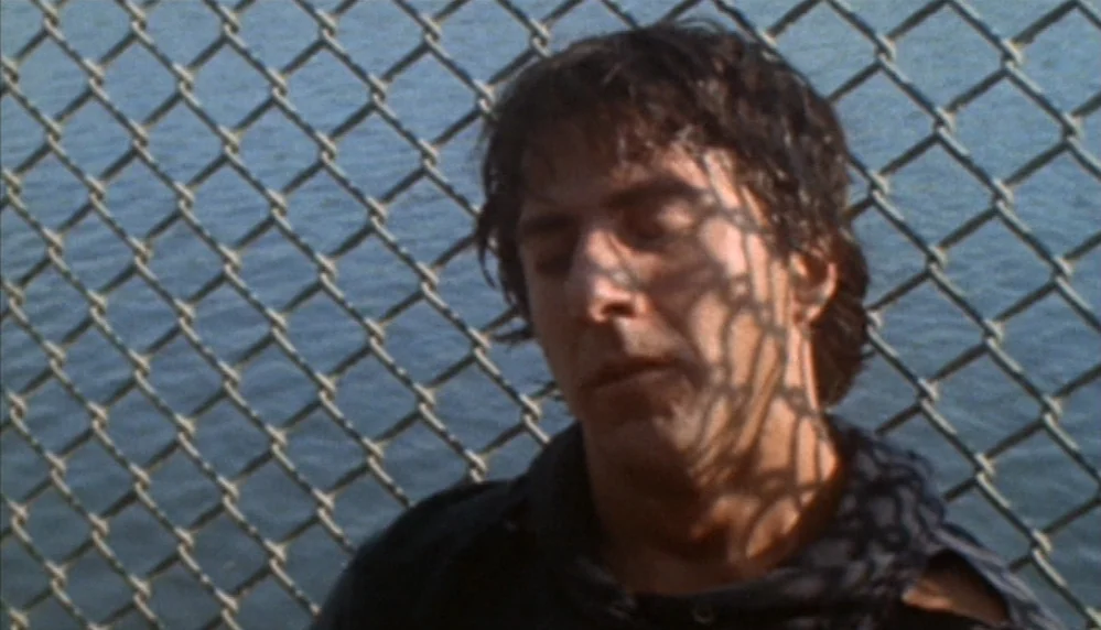 Marathon Man (1976) movie still: closeup — A man stands outdoors with a chain-link fence immediately behind him. The man has his eyes closed and…