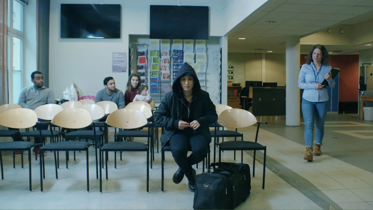 Code of Trust (2019) movie still: medium — A sterile public waiting area with rows of chairs and a bulletin board background. A young woman in a…