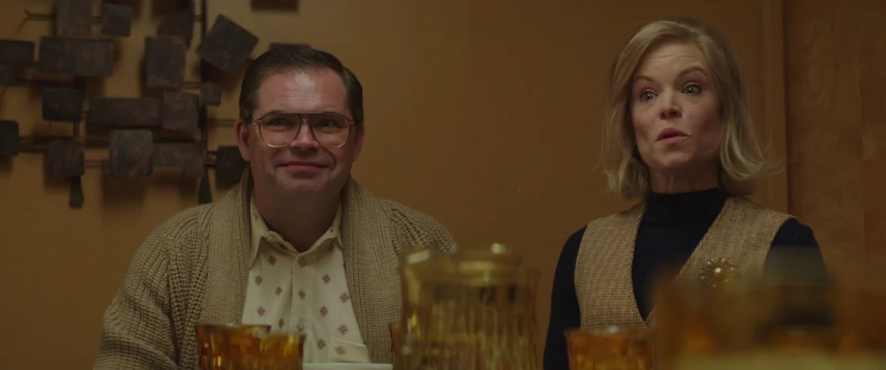 Southbound (2015) movie still: medium — A 1970s style dining room with golden-yellow walls and a decorative metal wall sculpture. A man in a…