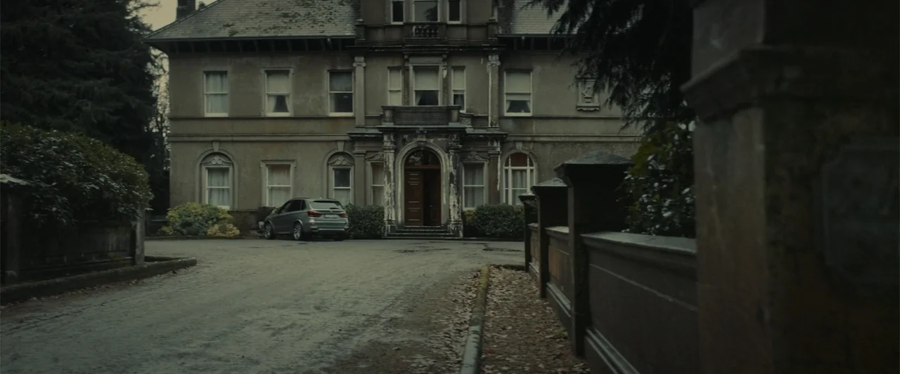 The Cellar (2022) movie still: establishing — A stone mansion with an arched doorway sits behind a curved stone wall. A silver SUV is parked on the…