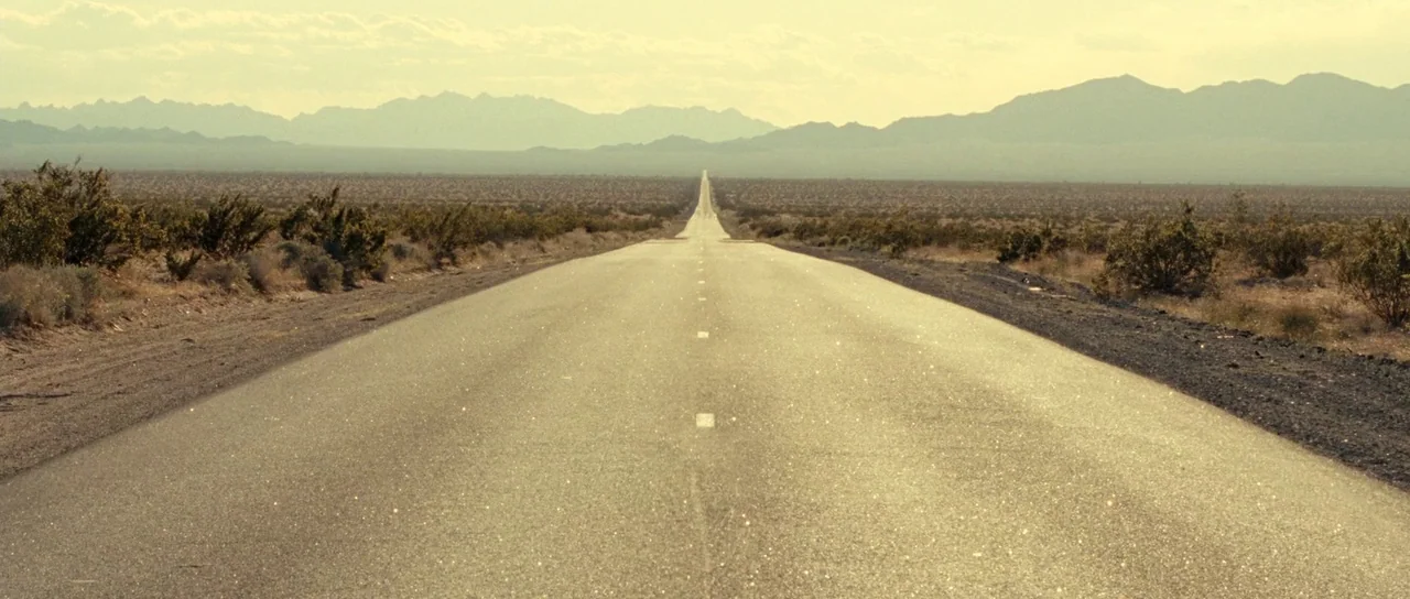 On The Road (2012) movie still: establishing — A long, straight asphalt road bisects a flat, arid desert plain. Distant mountains frame the horizon…