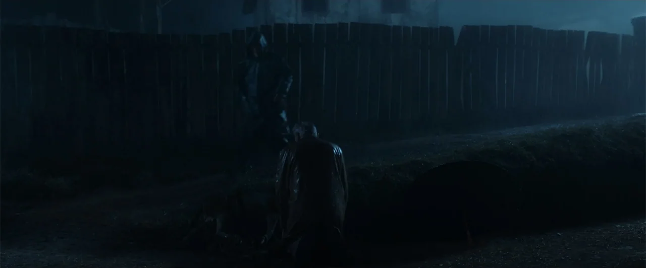 My Neighbor Adolf (2022) movie still: wide — A dark, wet dirt road at night is bordered by a tall wooden fence. A man kneels on the wet ground while…