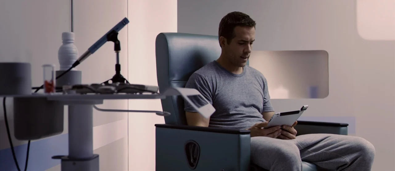 Self/less (2015) movie still: medium — A sterile medical room features smooth white walls and a specialized treatment chair. A man sits in the chair…