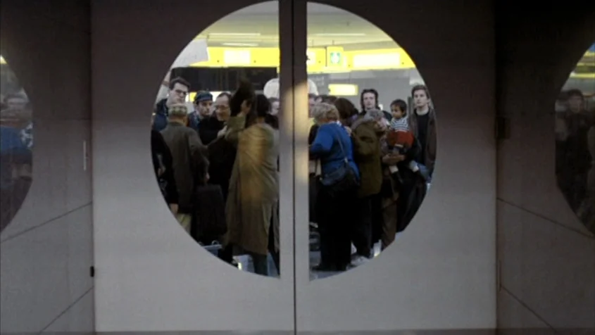 Benny’s Video (1992) movie still: medium — A group of people stands in a brightly lit, sterile public terminal space. They are seen through a pair…