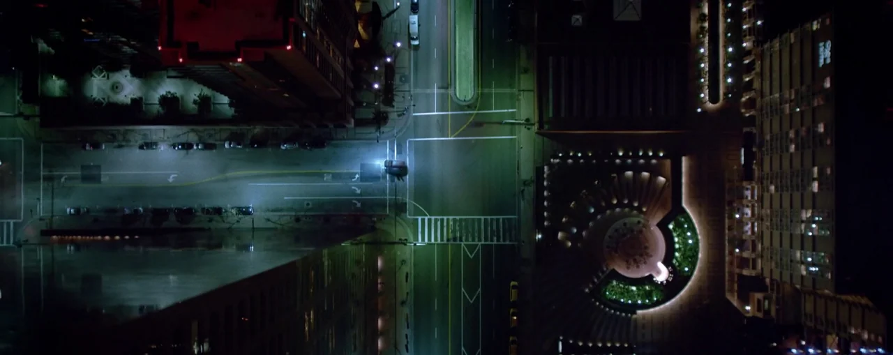 The Naked Gun (2025) movie still: establishing — A top-down view of a dark city intersection illuminated by overhead streetlights at night. A lone…