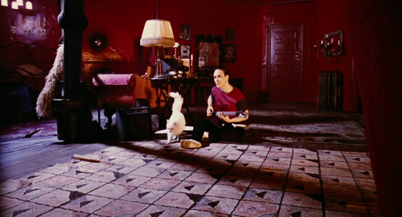 The Crow (1994) movie still: wide — A dark apartment features deep red walls and a large patterned carpet on floor. A man sits on the floor playing…