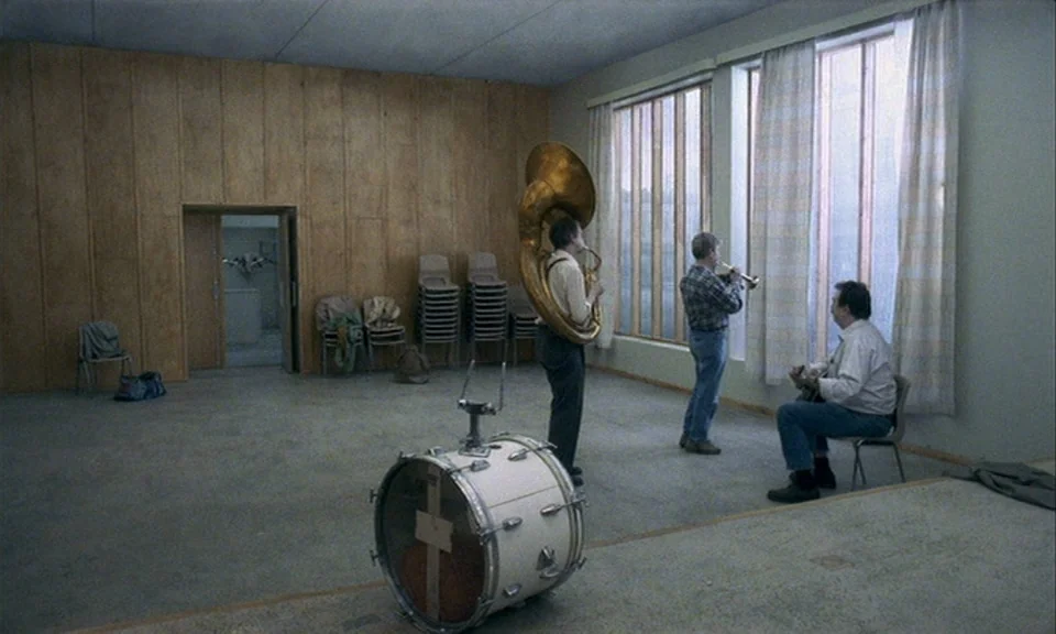 You, The Living (2007) movie still: wide — A sparsely furnished hall features wood-paneled walls and floor-to-ceiling windows with thin curtains.…