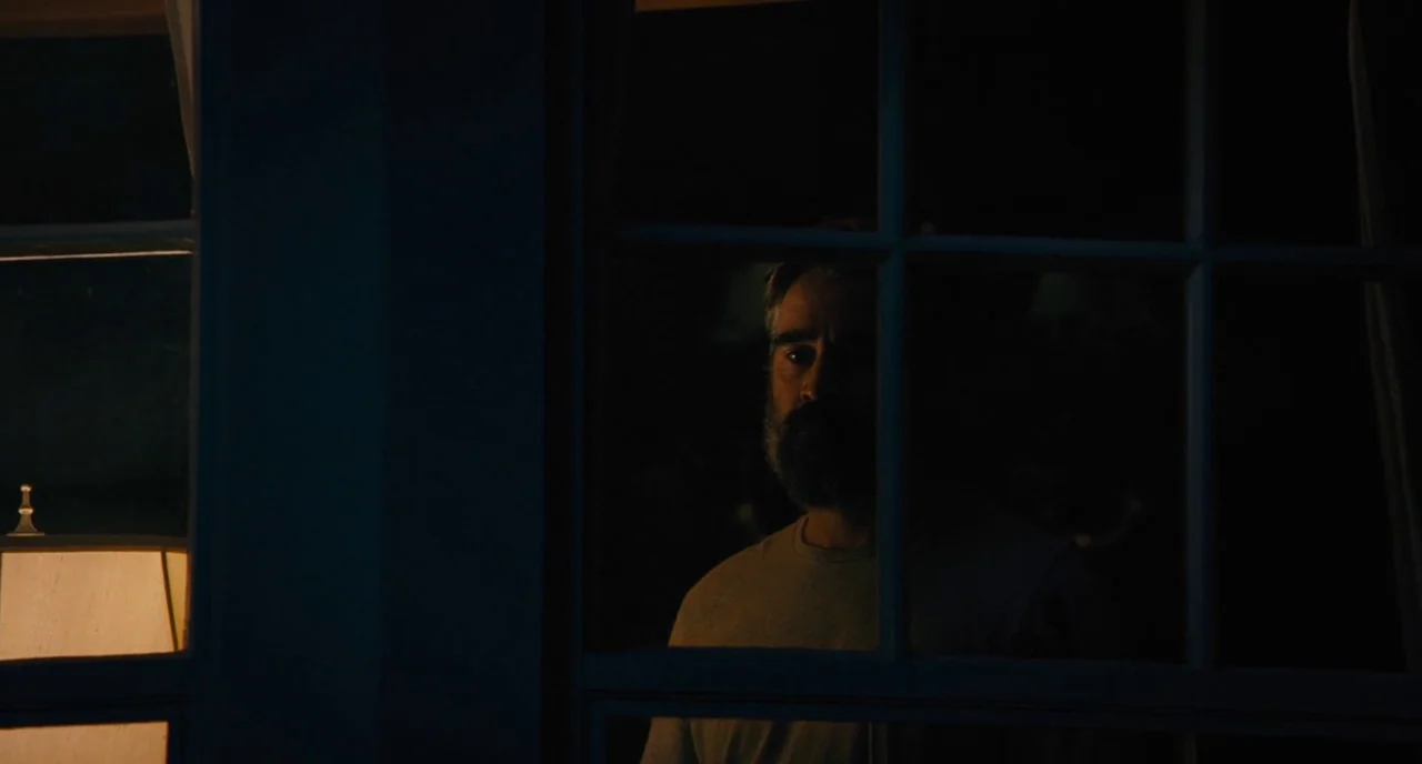 The Killing of a Sacred Deer (2017) movie still: medium — A dark, exterior view through a window pane into a dimly lit space. A man stands partially…
