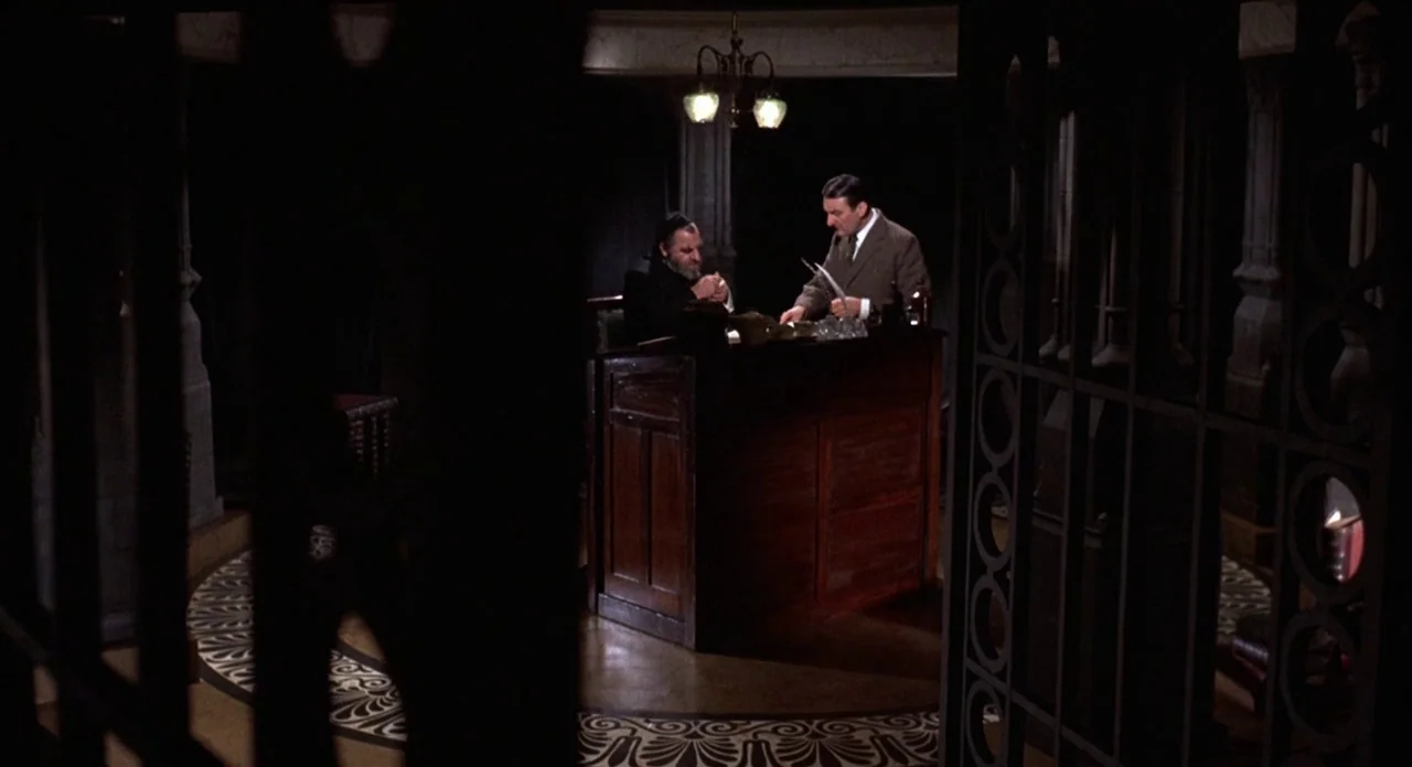 The Abominible Dr. Phibes (1972) movie still: wide — A dimly lit room features circular mosaic flooring and ornate metal gate dividers. Two men…