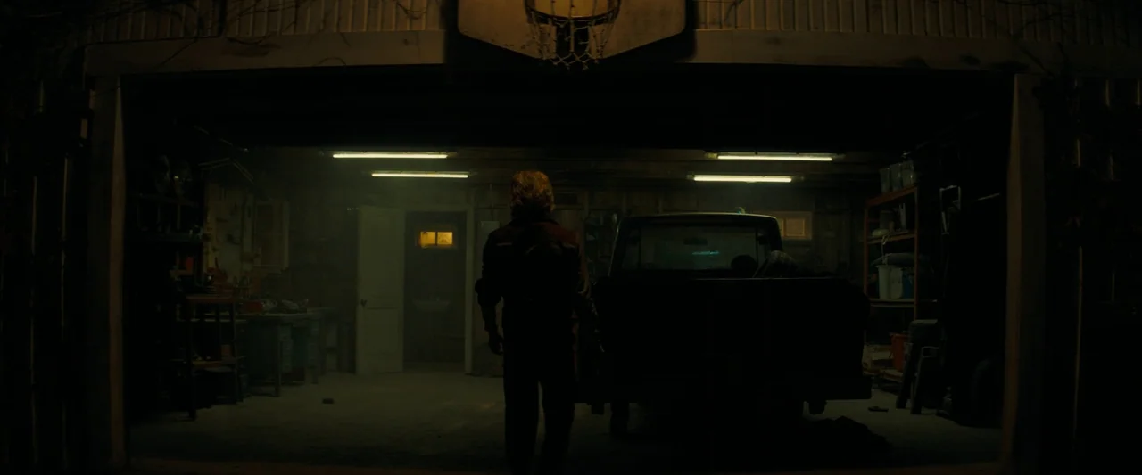 Don’t Breathe 2 (2021) movie still: fullbody — A cluttered, dimly lit garage interior contains a parked pickup truck and workbench shelves. A person…