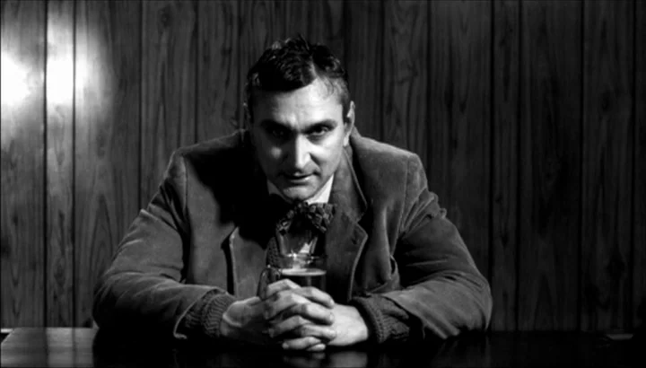 The American Astronaut (2001) movie still: medium — A dark space features a vertical wood-paneled wall behind a man sitting at a table. The man sits…
