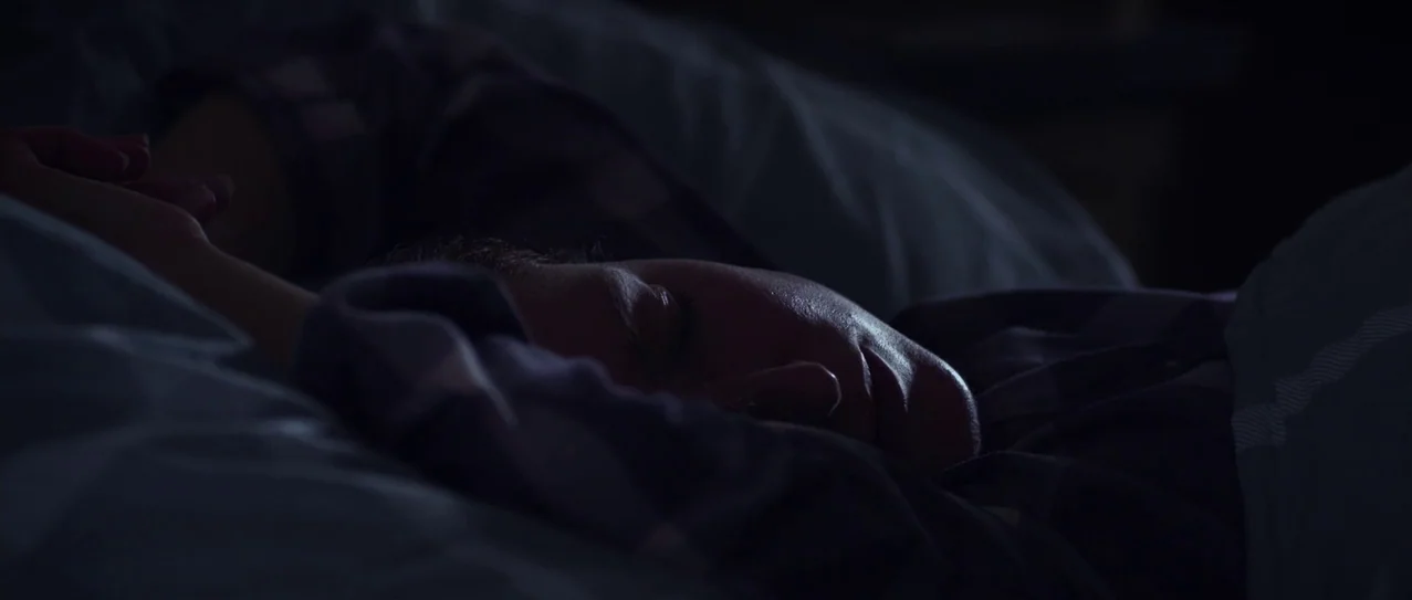 Vanishing Waves (2012) movie still: closeup — A person sleeps in a darkened room under rumpled bedding. The subject lies on their side with one arm…
