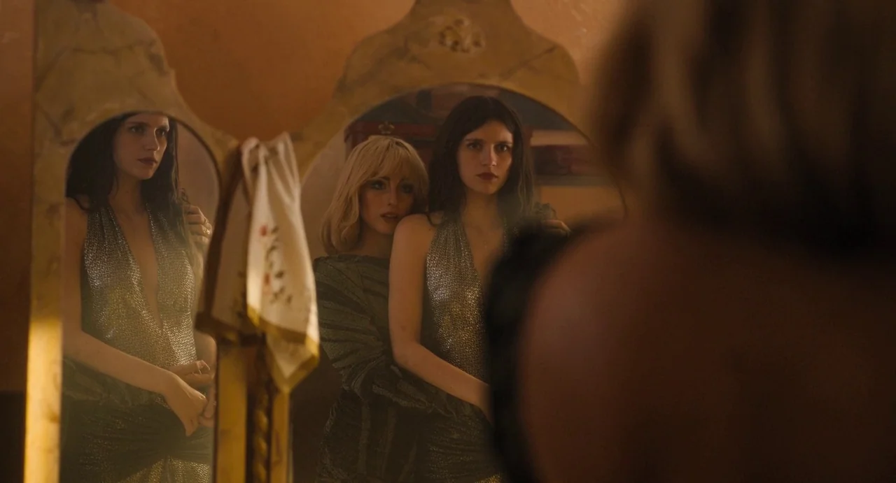 The First Omen (2024) movie still: medium — A dimly lit room contains an ornate antique mirror reflecting two women. One woman with blonde hair…
