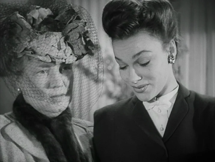 Crisis (1946) movie still: medium — An interior space features an older woman and a younger woman standing together. They look down at an object…