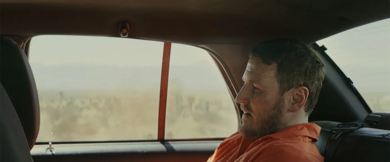 One Ranger (2023) movie still: medium — A man wearing an orange jumpsuit sits in the backseat of a moving car. Outside the window, a blurred desert…