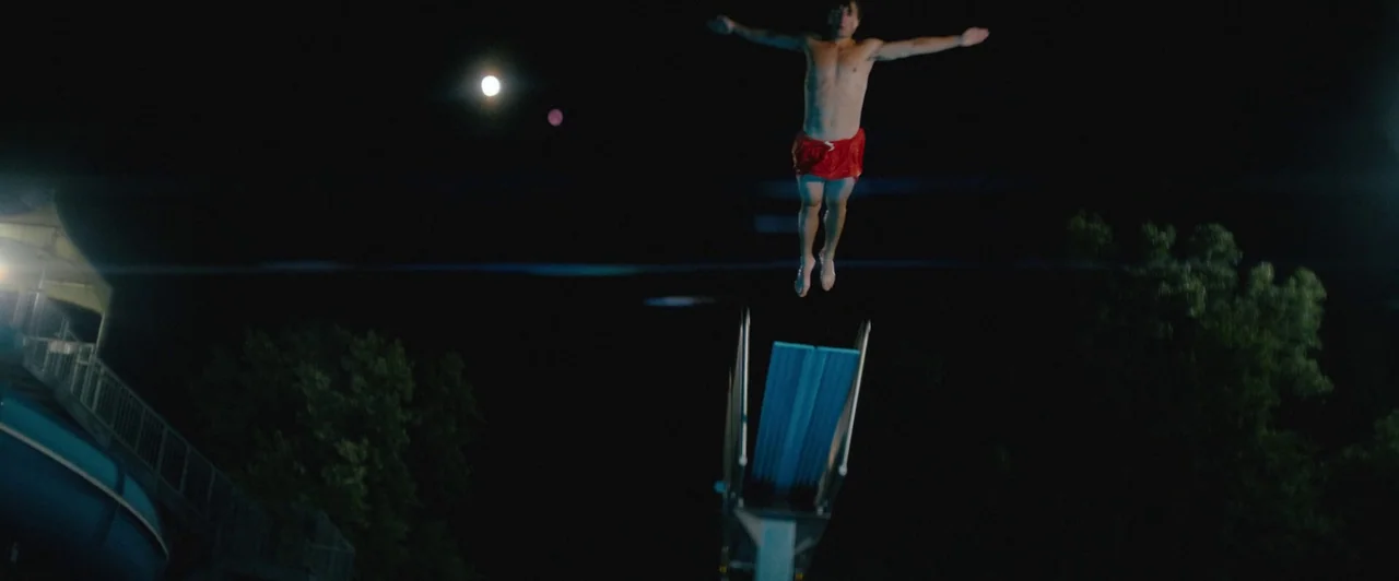 Snack Shack (2024) movie still: wide — A night view at a pool with a lone figure in mid-air. The man jumps from a blue diving board into the darkness.
